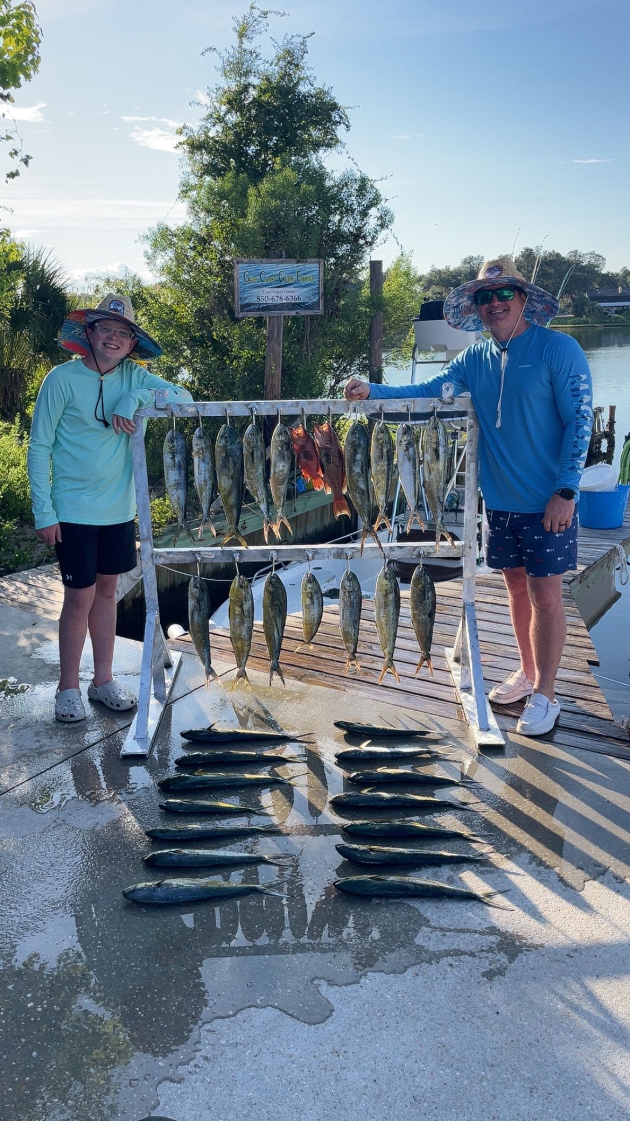 Two people display a large catch of fish on a rack and ground, outdoors by water.