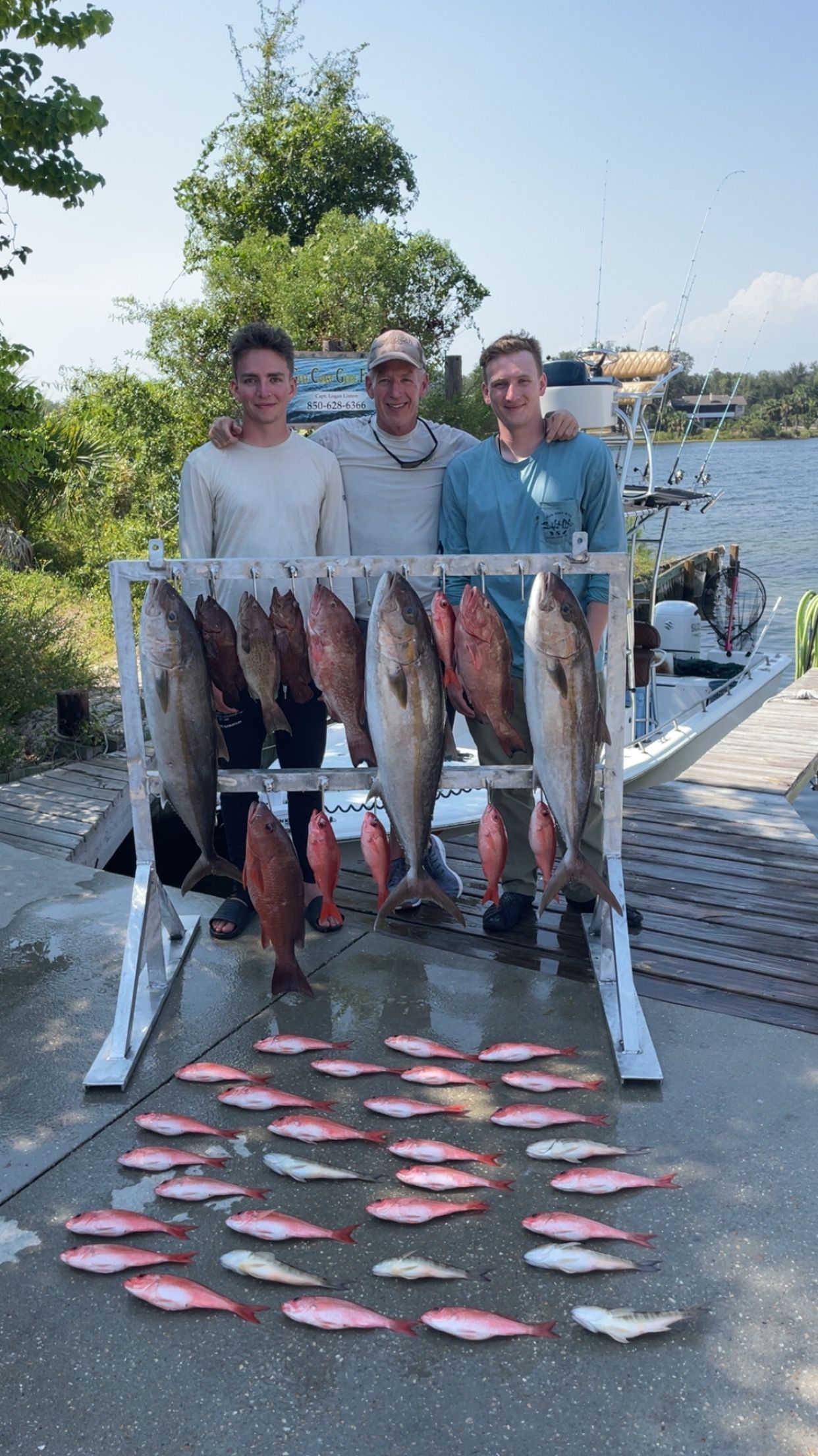 Three men pose with a large fish catch on a dock. Red and larger gray fish hang on a rack and are piled below.