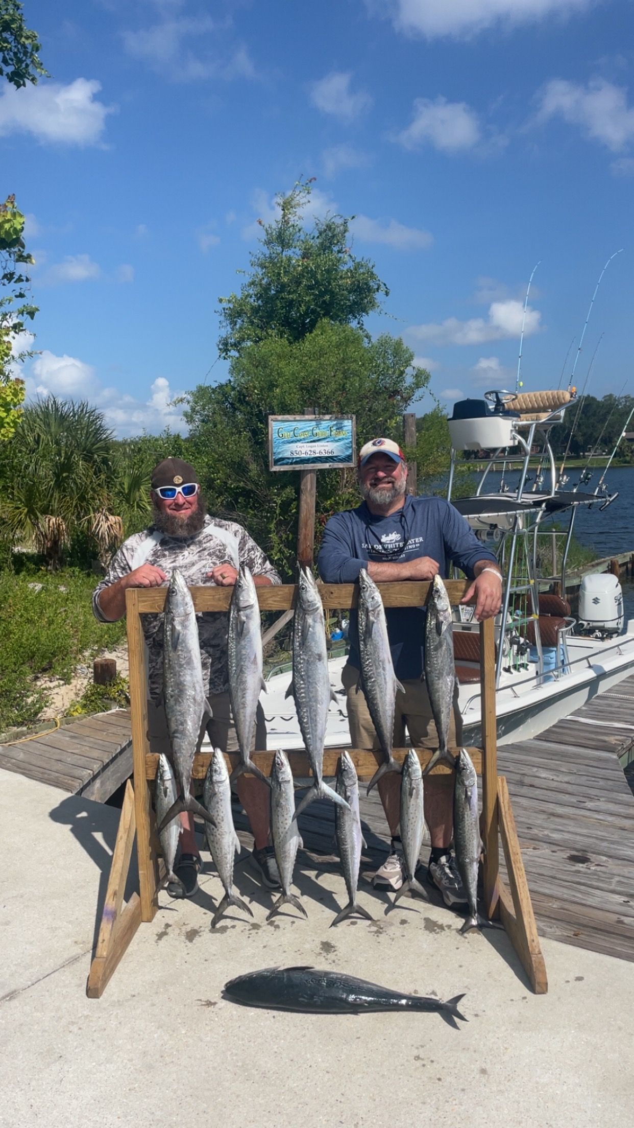Two men with fishing catch, posing outdoors near a dock. Silver fish displayed on a wooden rack, sunny day.