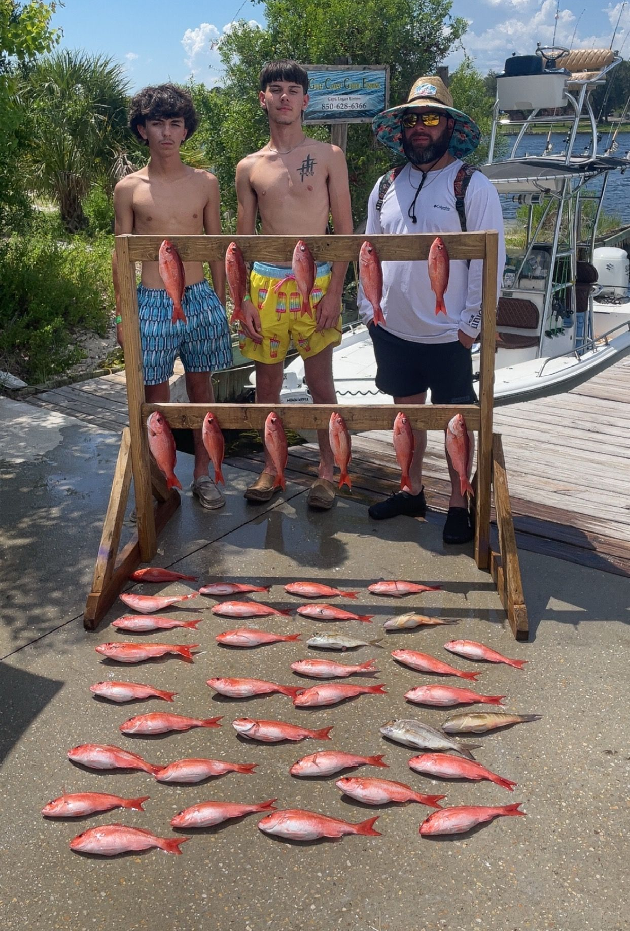 Three people pose with a display of caught red fish near a dock on a sunny day.
