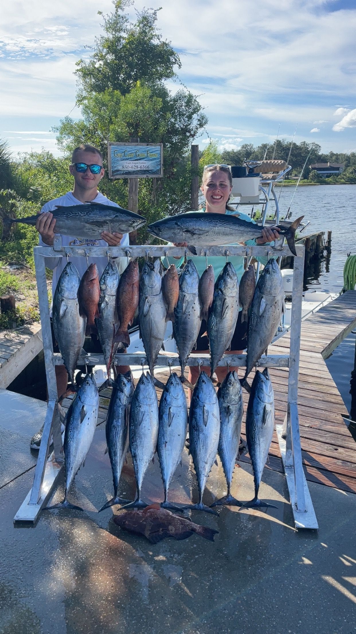 Two people display many fish on a rack near water. Clear skies, green trees, and a sign are visible.
