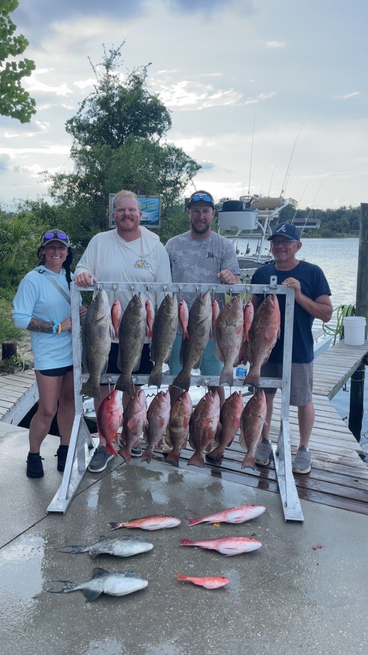 Four people pose with a large fish catch displayed on a rack at a dock, including red snapper and grouper.