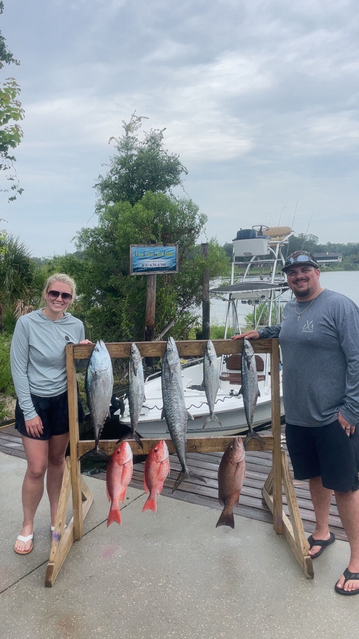 Two people pose with a fishing catch, showing various fish suspended from a wooden rack near a body of water.