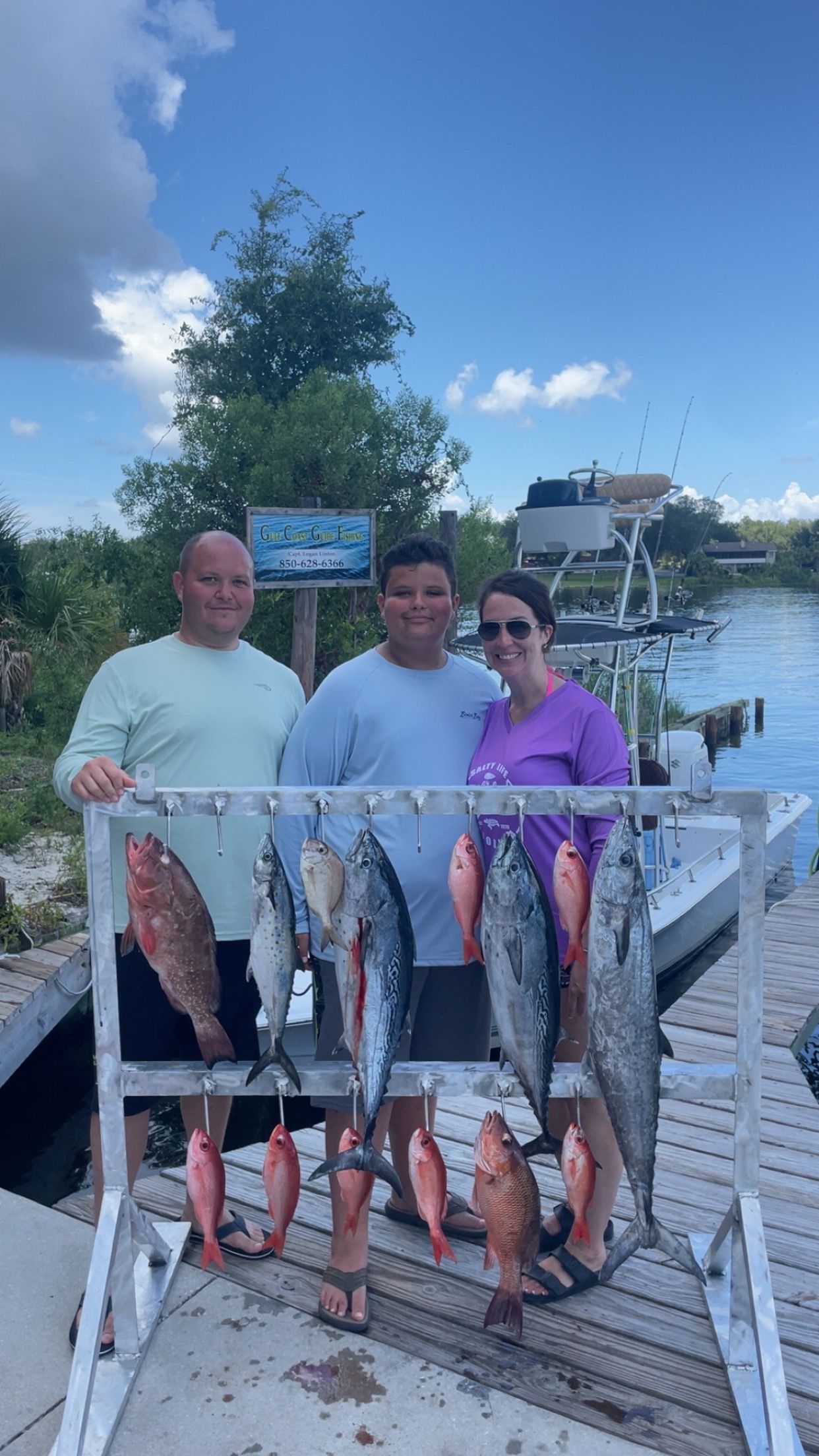 Three people smiling with a rack of various fish on a dock. Sky and water background.