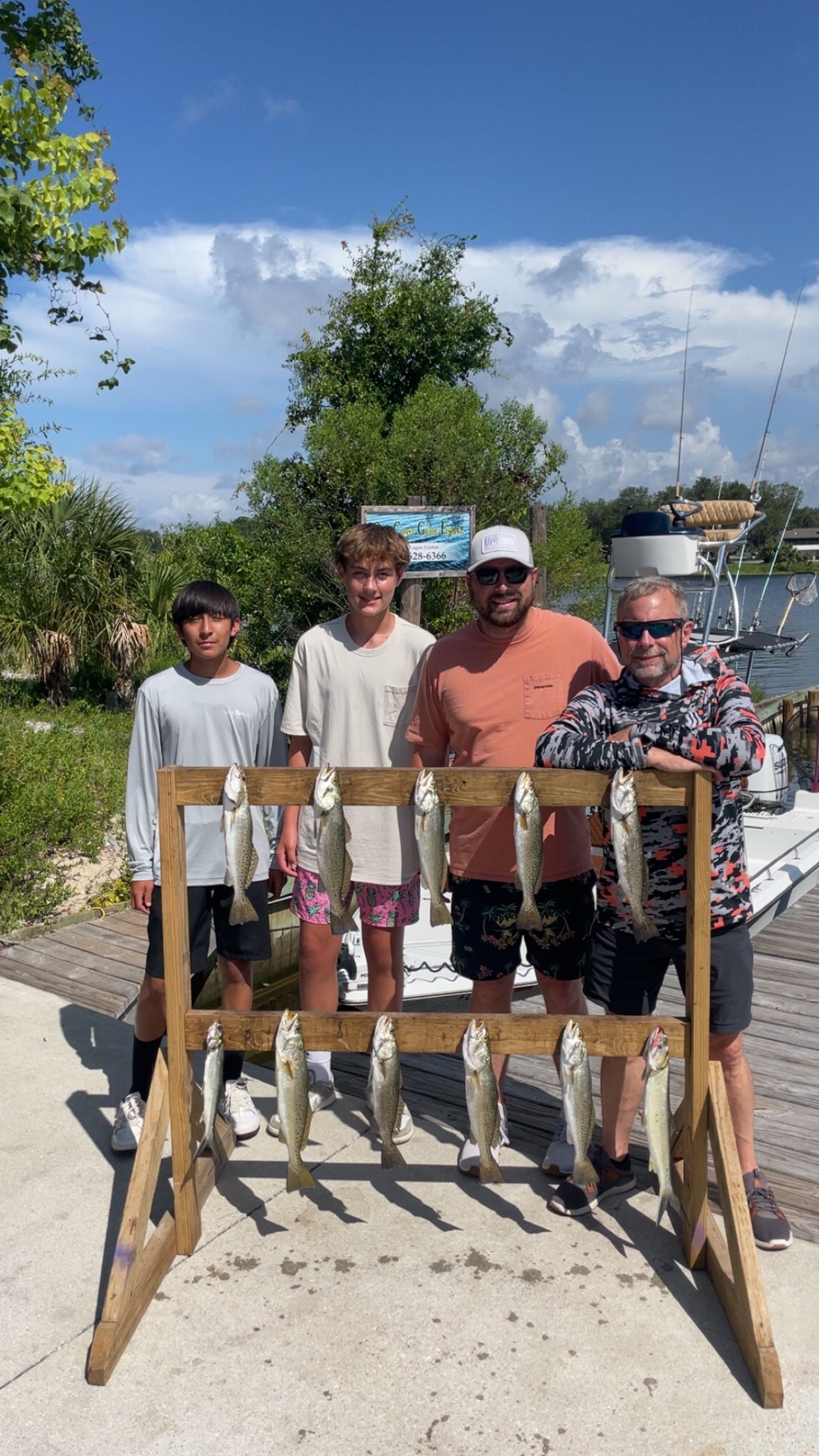 Four people pose with a wooden rack displaying several fish they caught. Outdoors, sunny day.