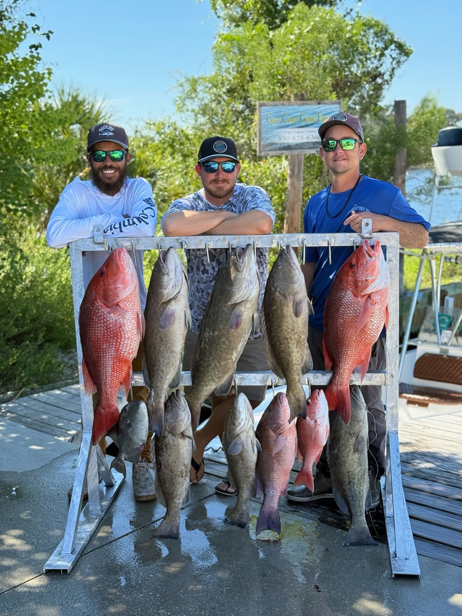 Three men pose with a fish rack full of caught fish outdoors near water.