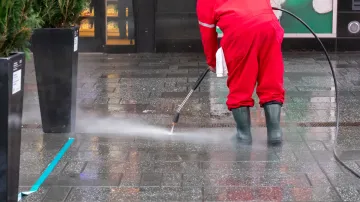 A man in a red jumpsuit is using a high pressure washer to clean a sidewalk.