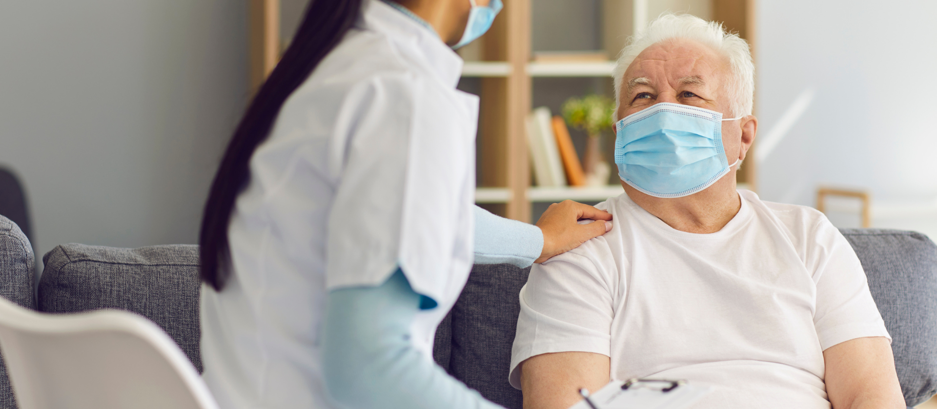 An elderly man wearing a mask is sitting on a couch with a nurse.