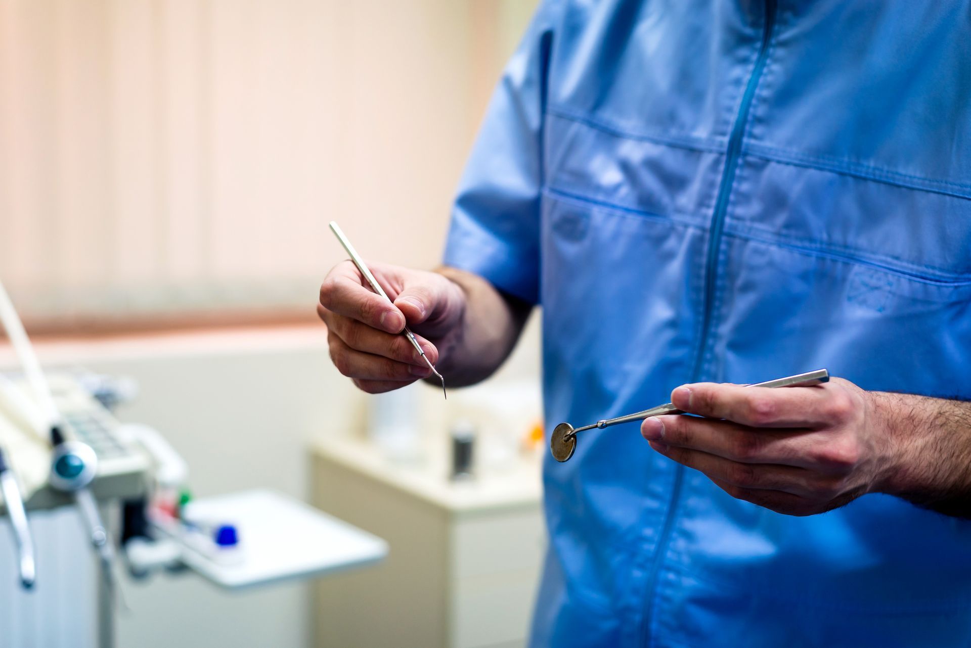Dentist in blue scrubs holding dental mirror and explorer tools in office. Dentist in blue scrubs holding dental mirror and explorer tools in office.