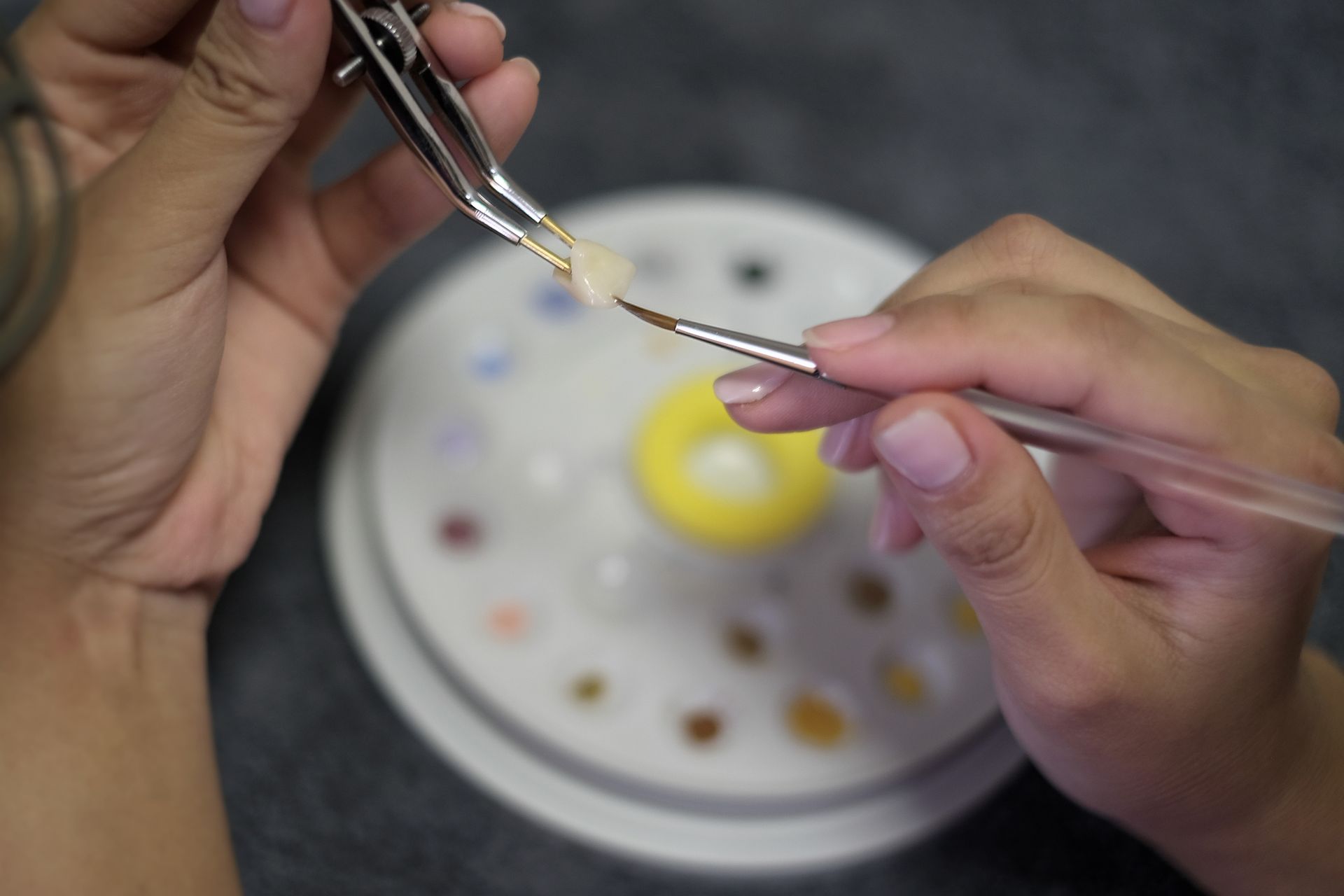 Hands painting a small object with a brush, held in tweezers, next to a palette of colors.