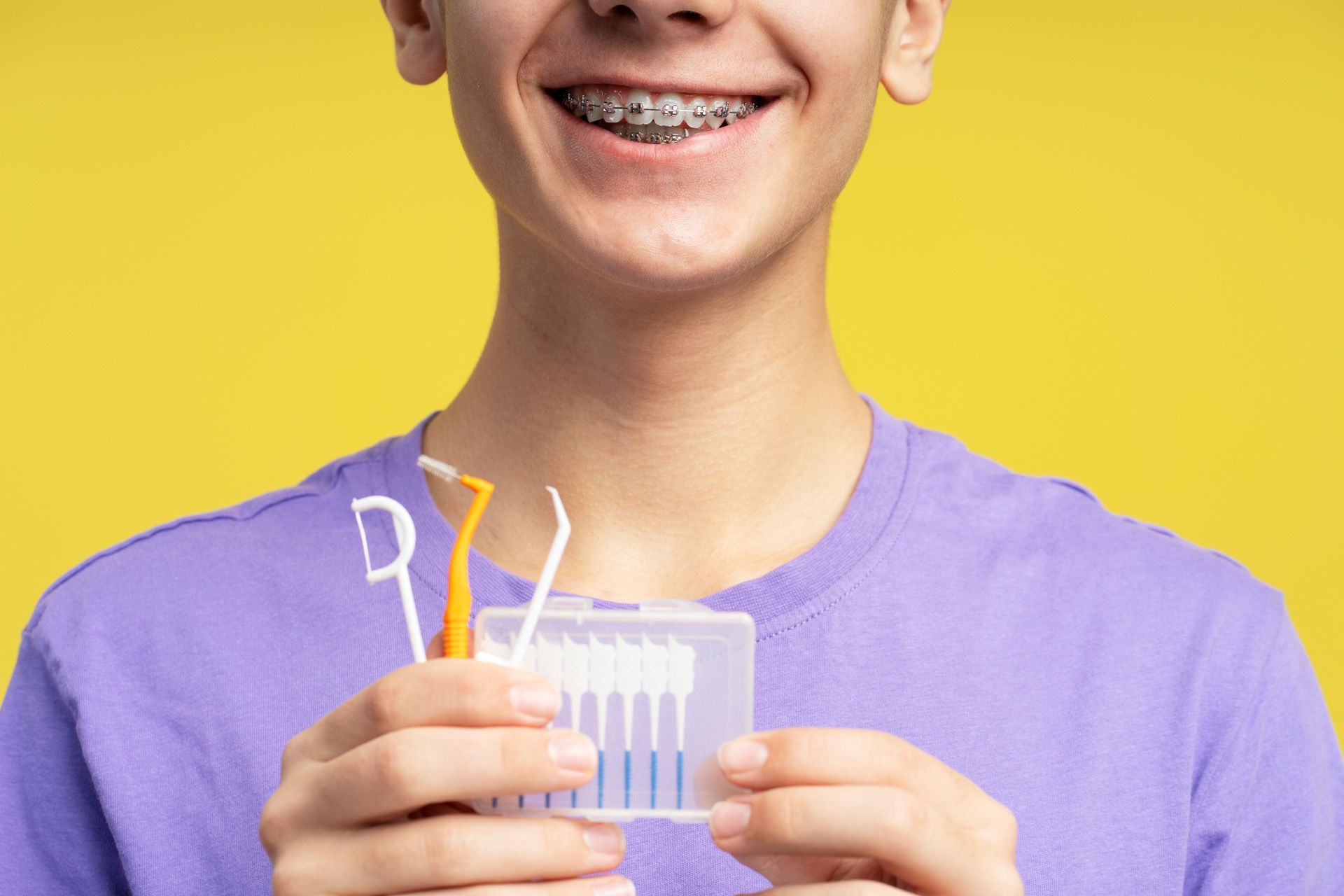 Person with braces holding dental cleaning tools, smiling against a yellow background.