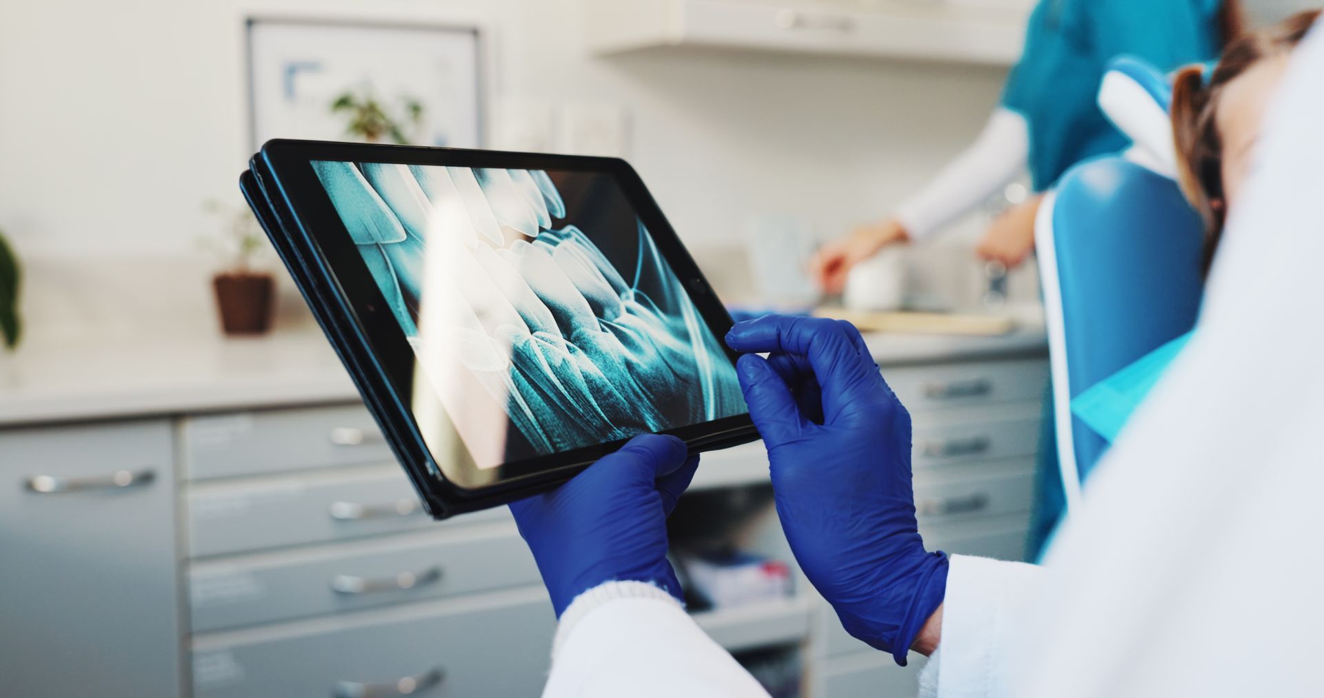 Dentist examining dental X-ray on a tablet in a clinic; blue gloves, white coat, dental chair. Dentist examining dental X-ray on a tablet in a clinic; blue gloves, white coat, dental chair.