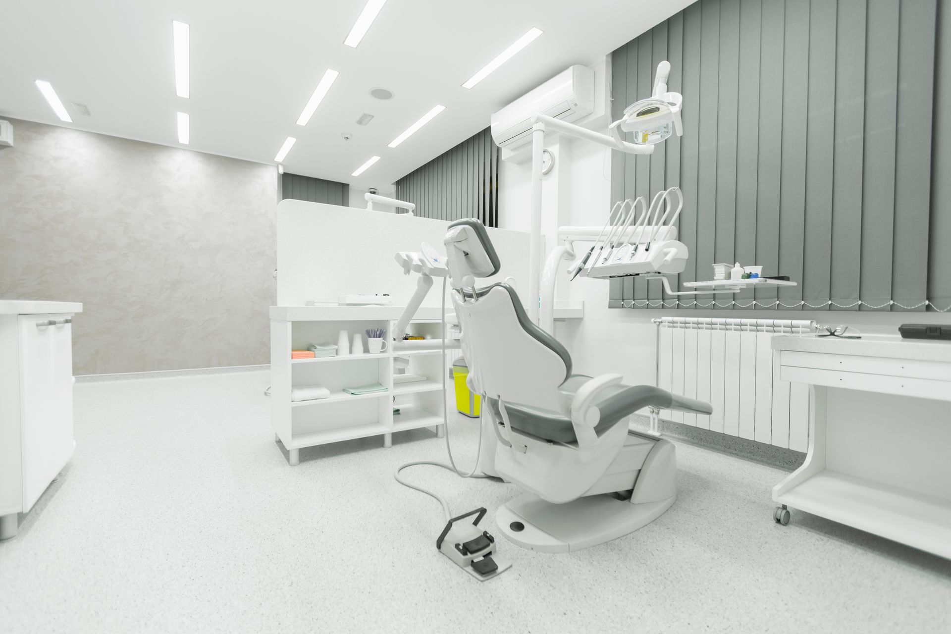 Bright, modern dentist's office with dental chair, equipment, cabinets, and window with blinds.