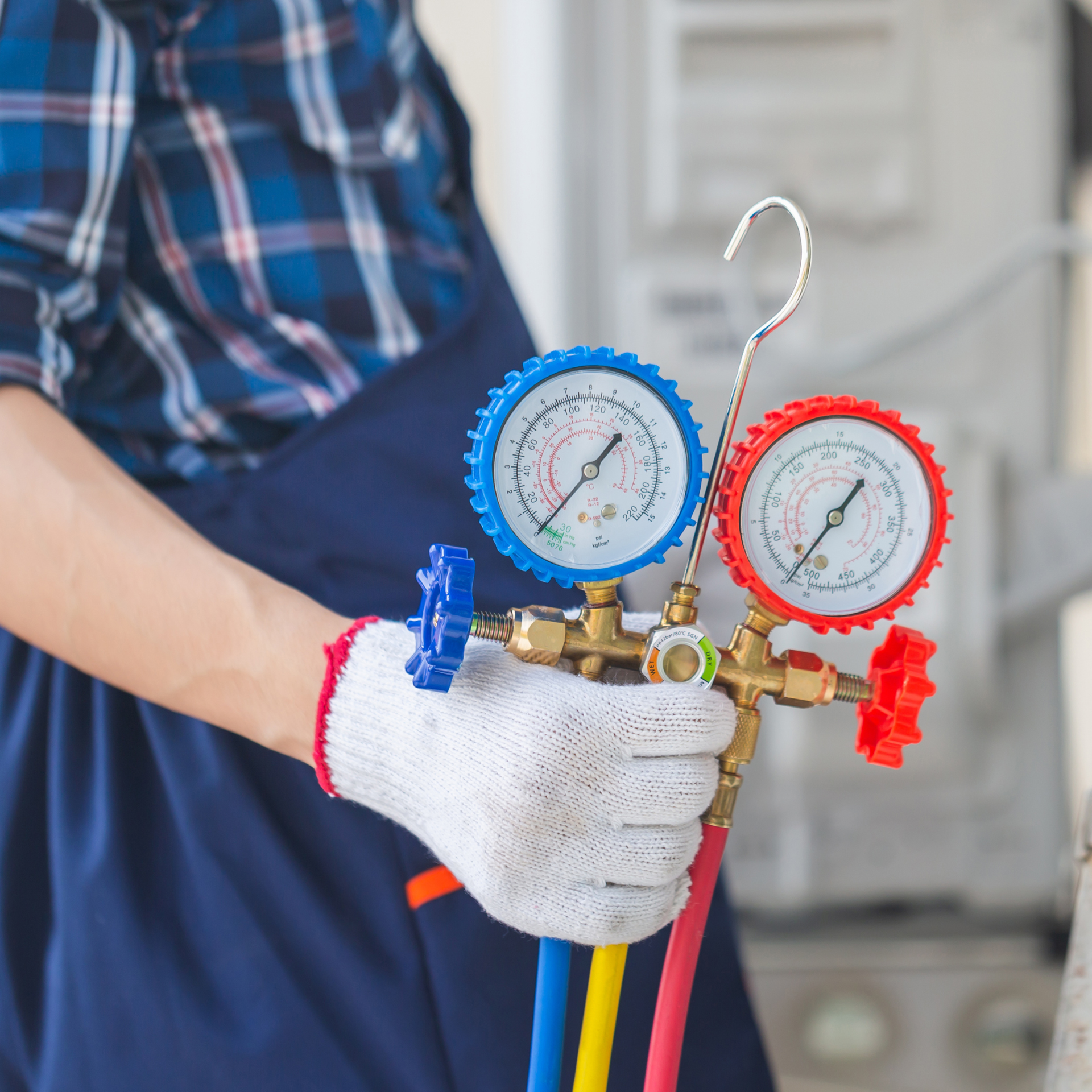 HVAC technician holding gauges, wearing gloves, in blue overalls, and a plaid shirt.