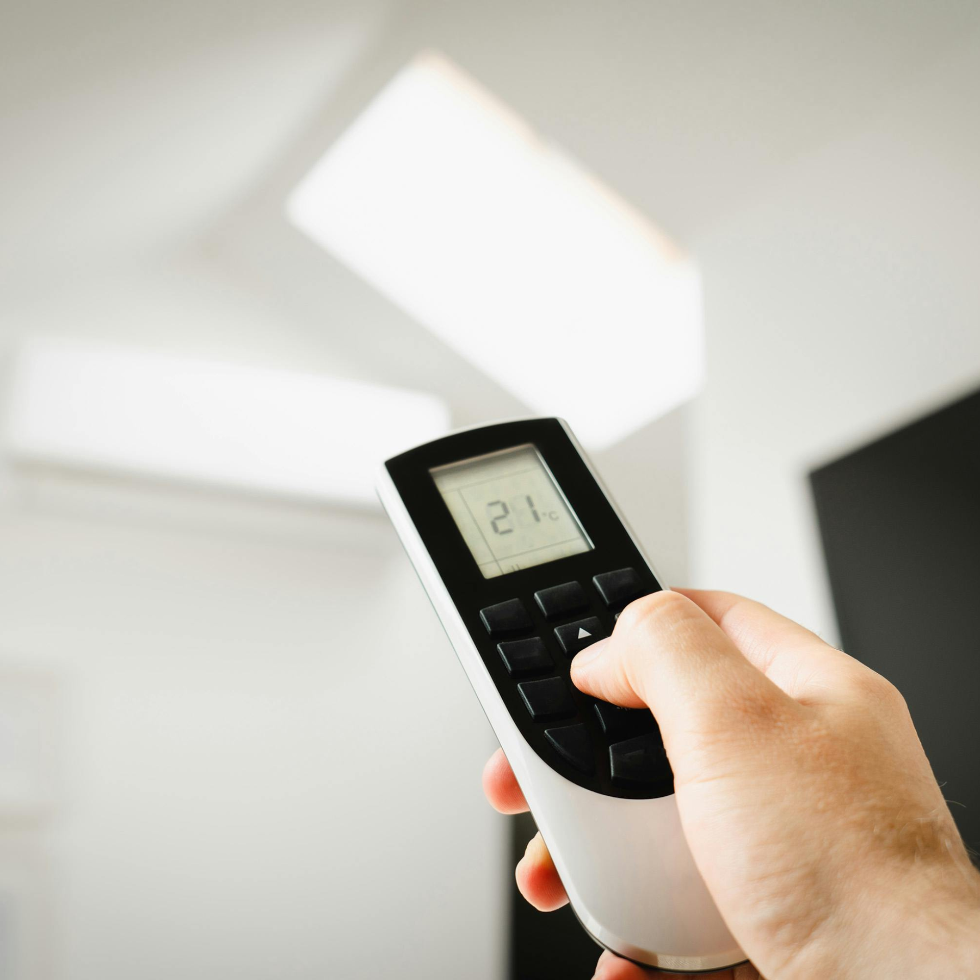 Hand holding a remote controlling an AC unit in a white room. Display reads 21.