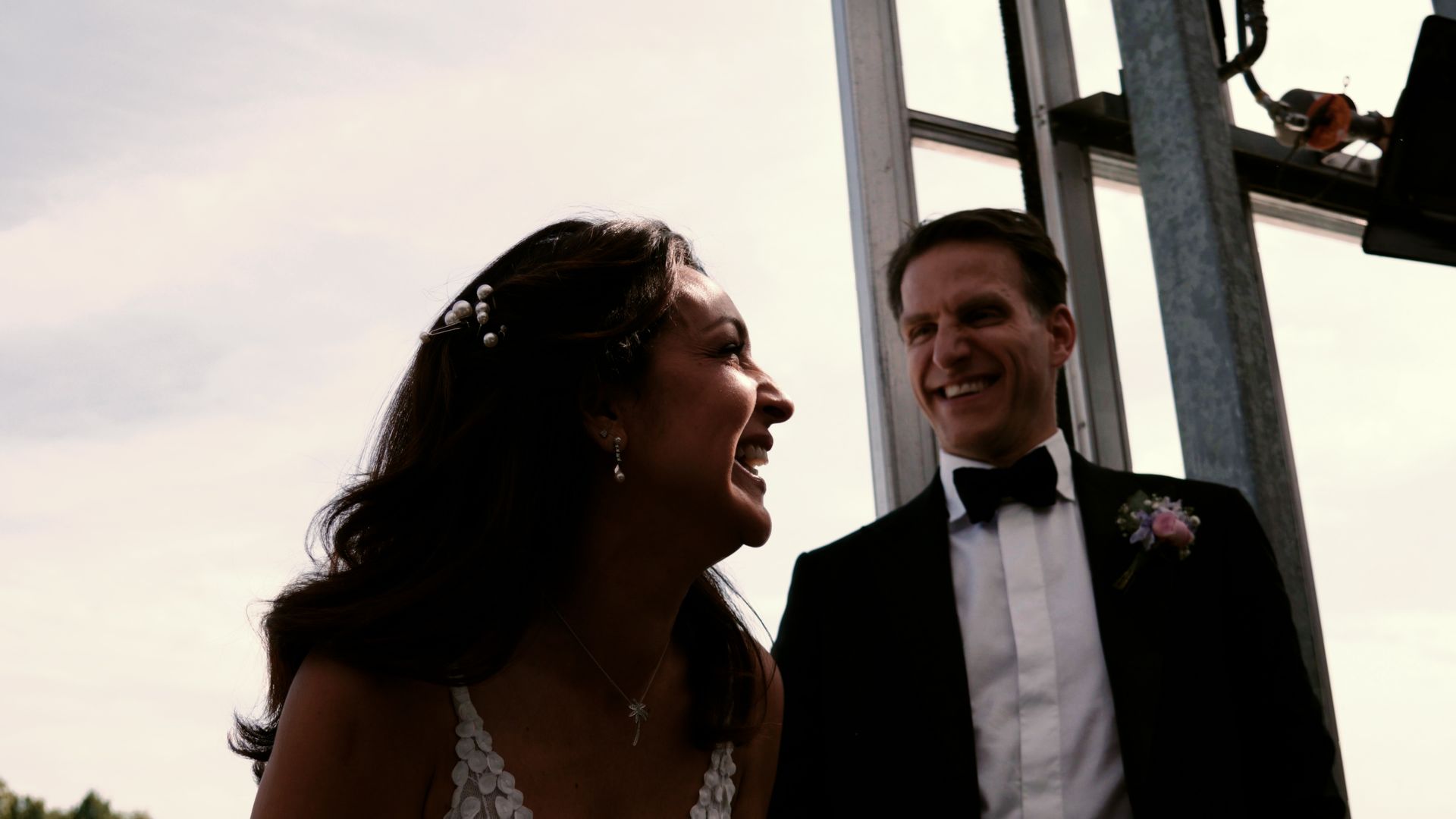 Two people in formalwear smiling outdoors at sunset, with a wedding dress visible on the left.