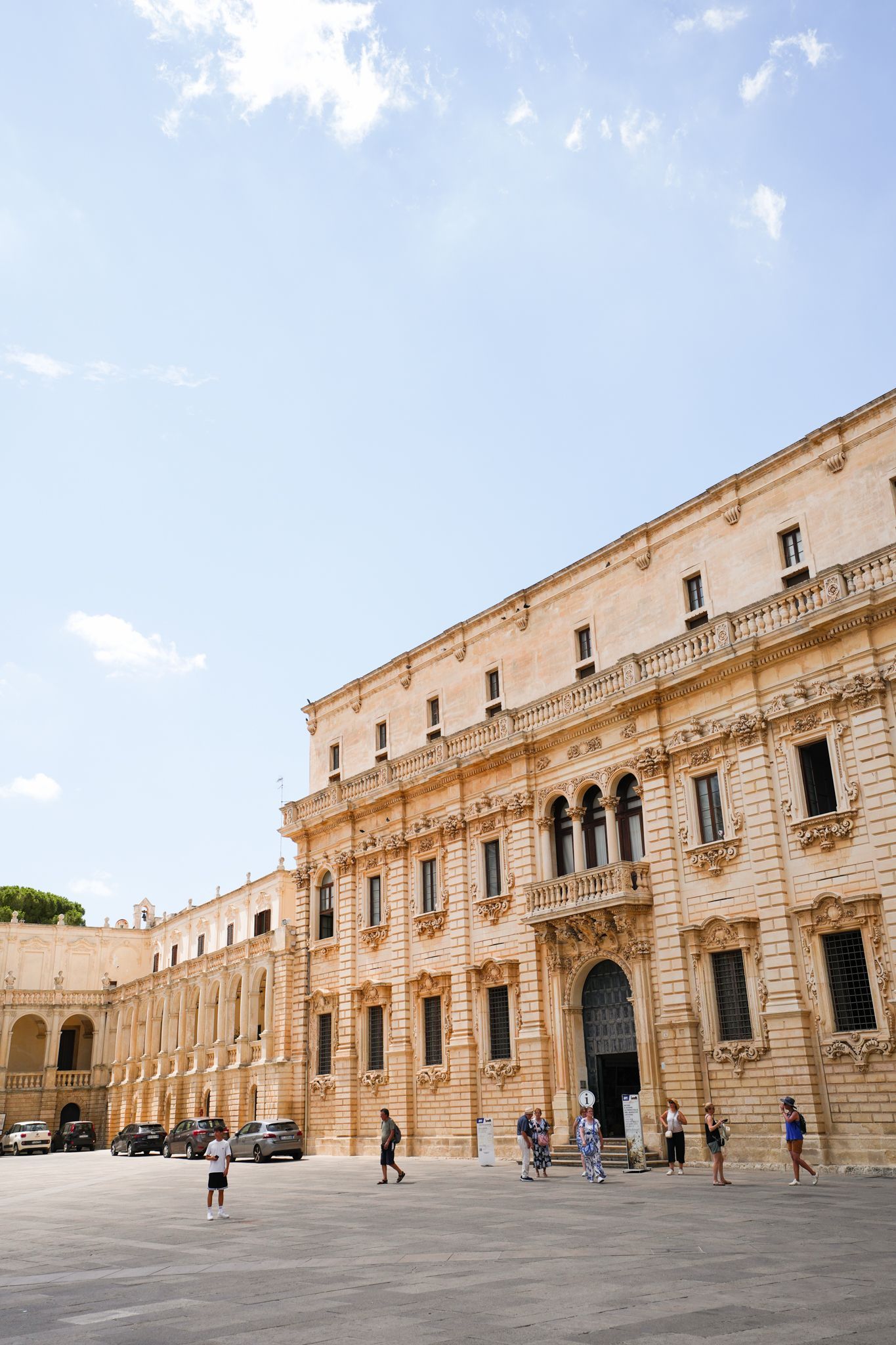 Historic stone building with arched windows and people in a sunlit courtyard under a blue sky