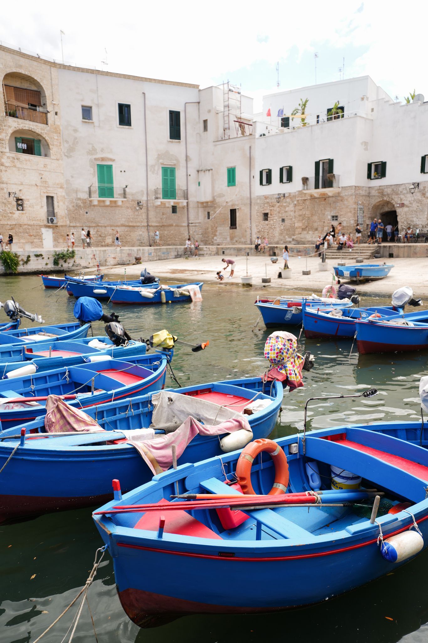Blue boats floating in a narrow harbor beside white waterfront buildings and a stone wall