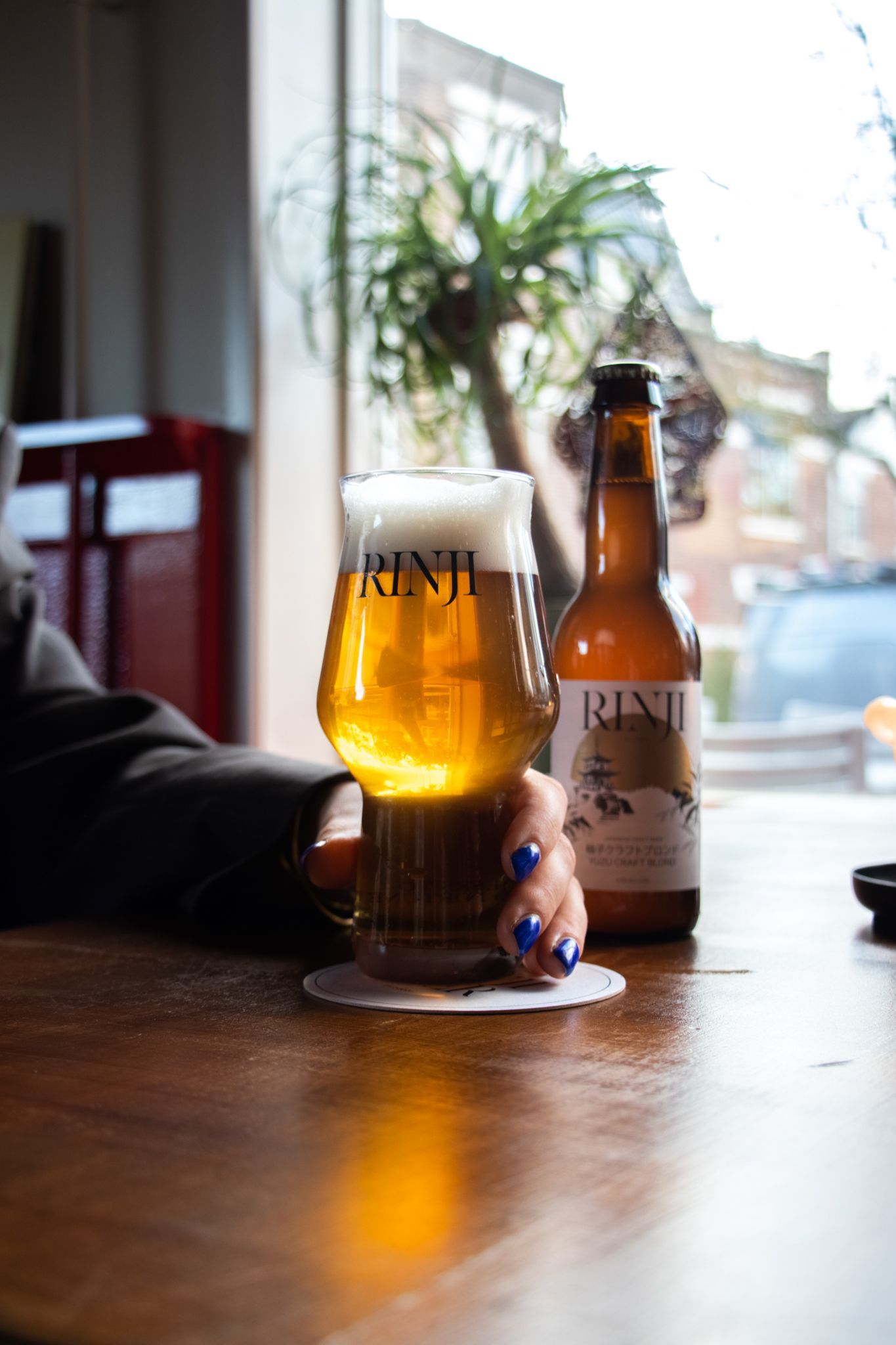 Beer glass and bottle on a wooden table by a window, with a blurred indoor background.