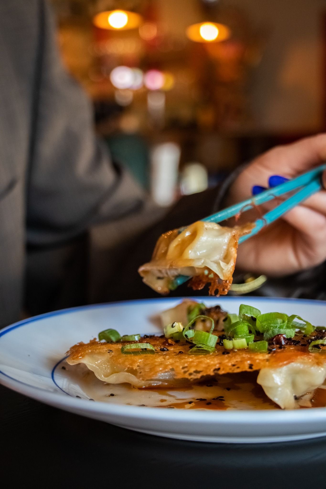 Hand holding chopsticks with food over a plated dish in a restaurant setting