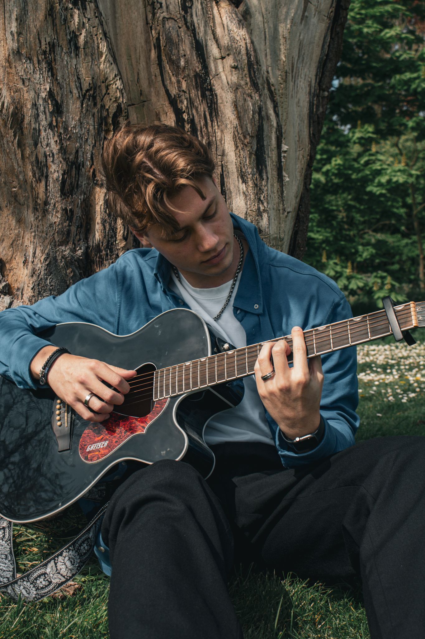 Een jongeman zit in het gras en bespeelt een akoestische gitaar.