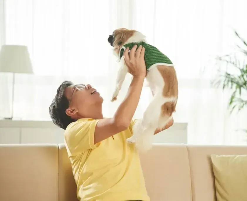 A man is sitting on a couch holding a dog in the air.