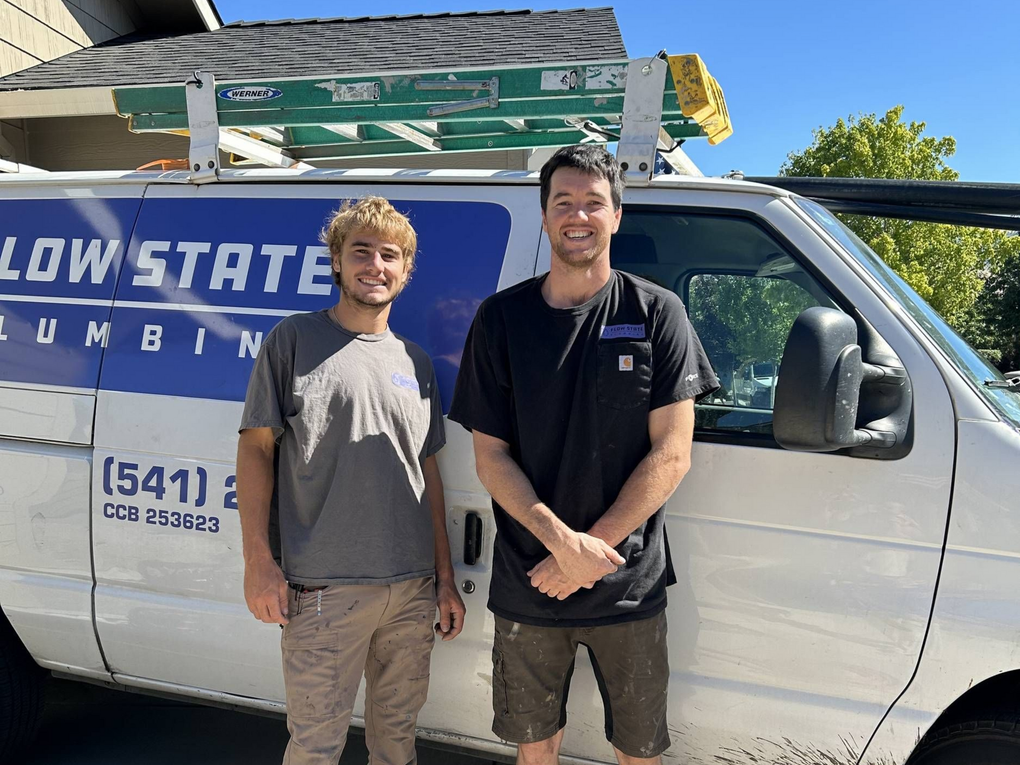 Two plumbers stand in front of a white van labeled 