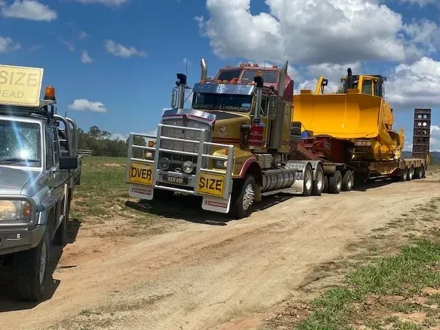 Yellow Truck Hauling a Large Yellow Bulldozer on a Trailer on a Dirt Road — Huntlys Heavy Equipment In Gracemere, QLD