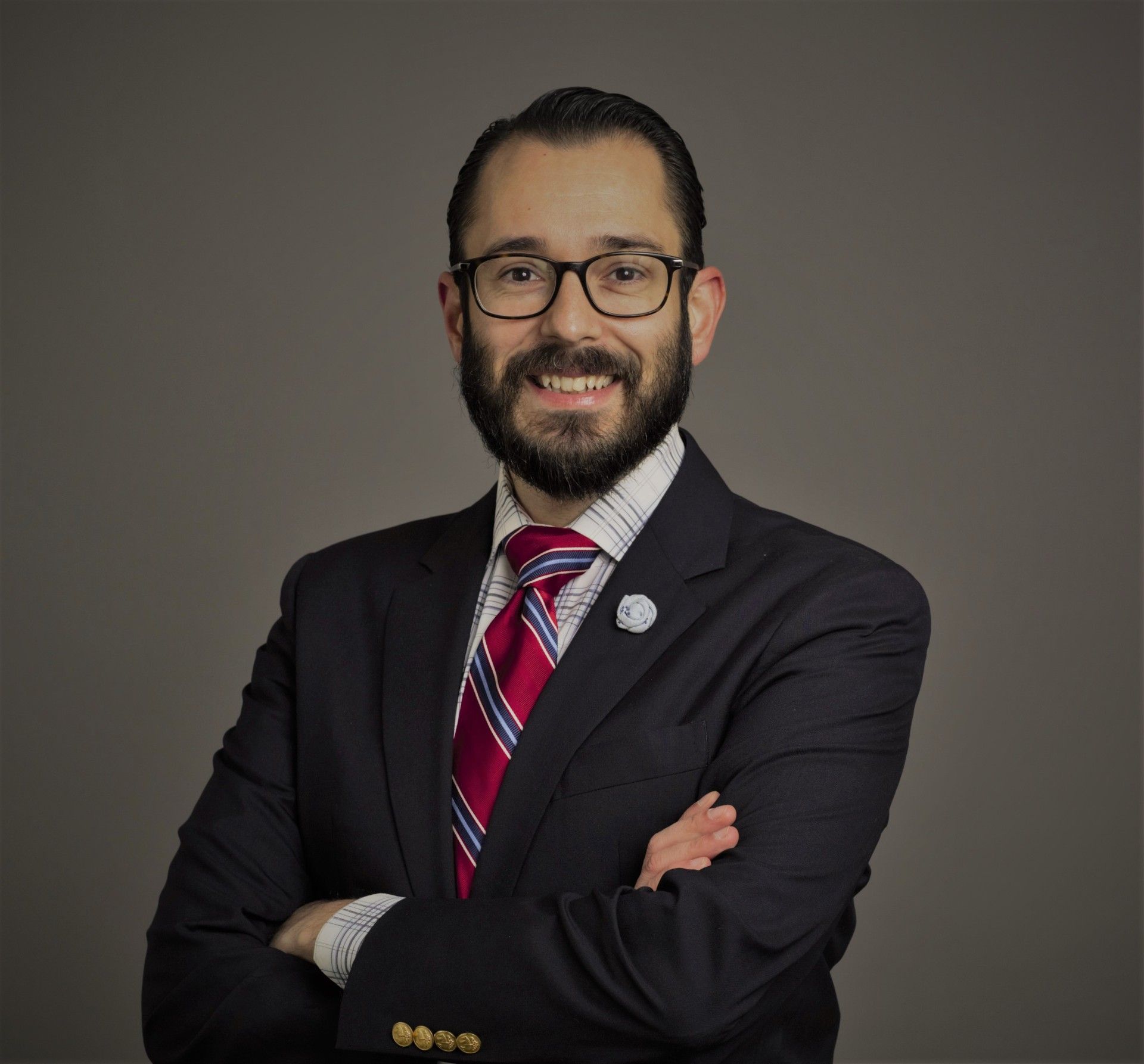 Man in a suit, arms crossed, smiling with glasses and a red and blue striped tie.