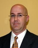 Bald man with glasses in a suit and tie, facing forward against a yellow wall.