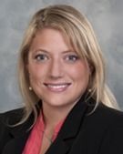 Blonde woman smiling, wearing a black blazer and pink shirt, against a gray background.