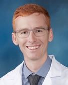 A fair-skinned man with red hair and glasses smiles, wearing a white coat and tie, likely a doctor.