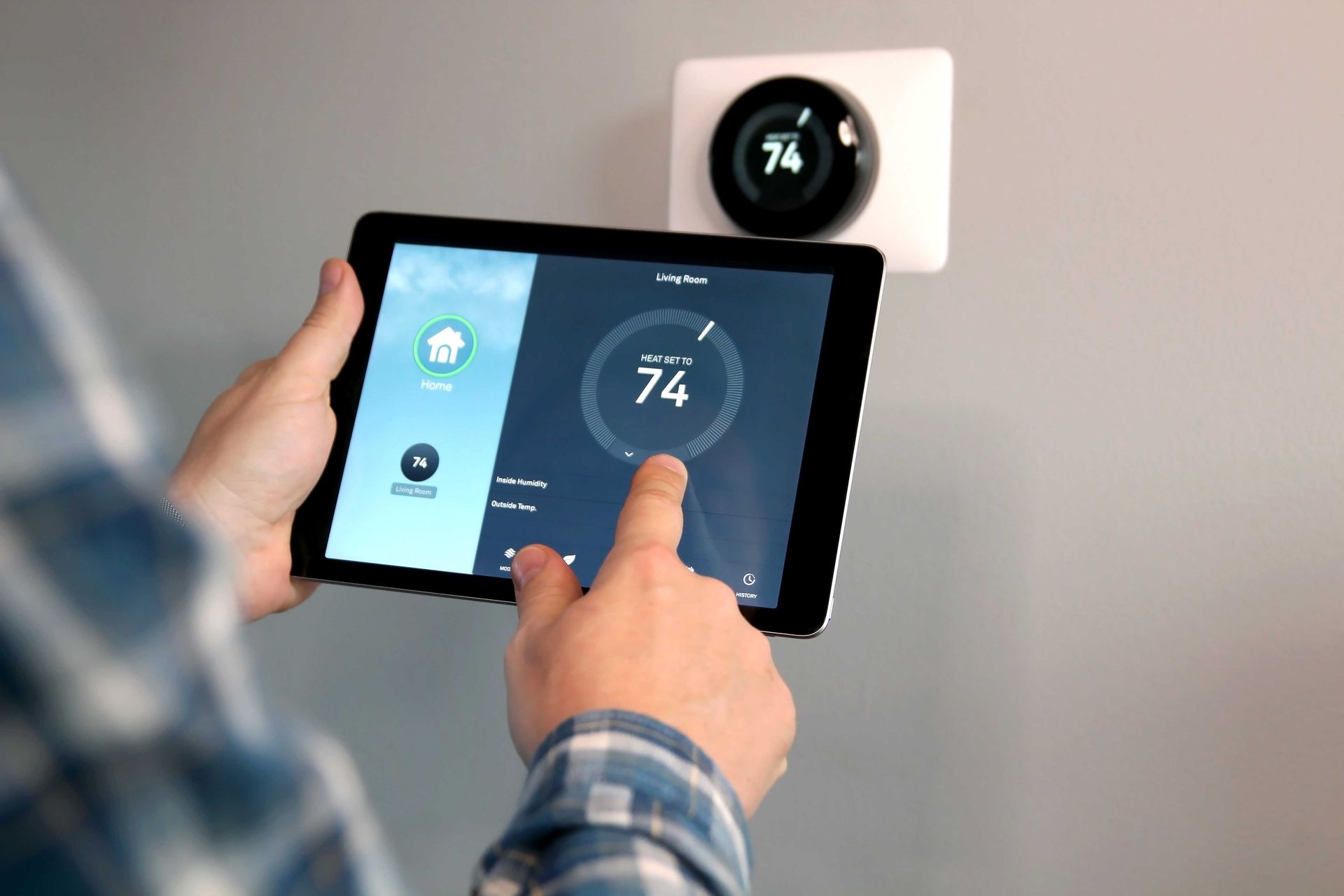 Hands adjusting a tablet thermostat showing 74°, with a wall-mounted smart thermostat behind it