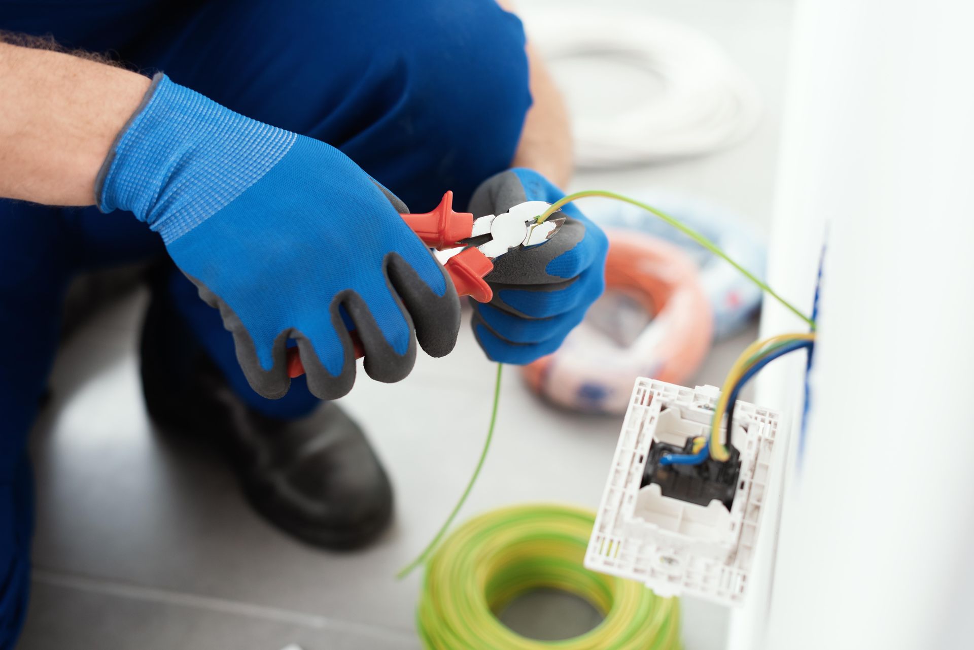 Gloved hands crimping a wire on an electrical cable beside tools and a wall outlet box