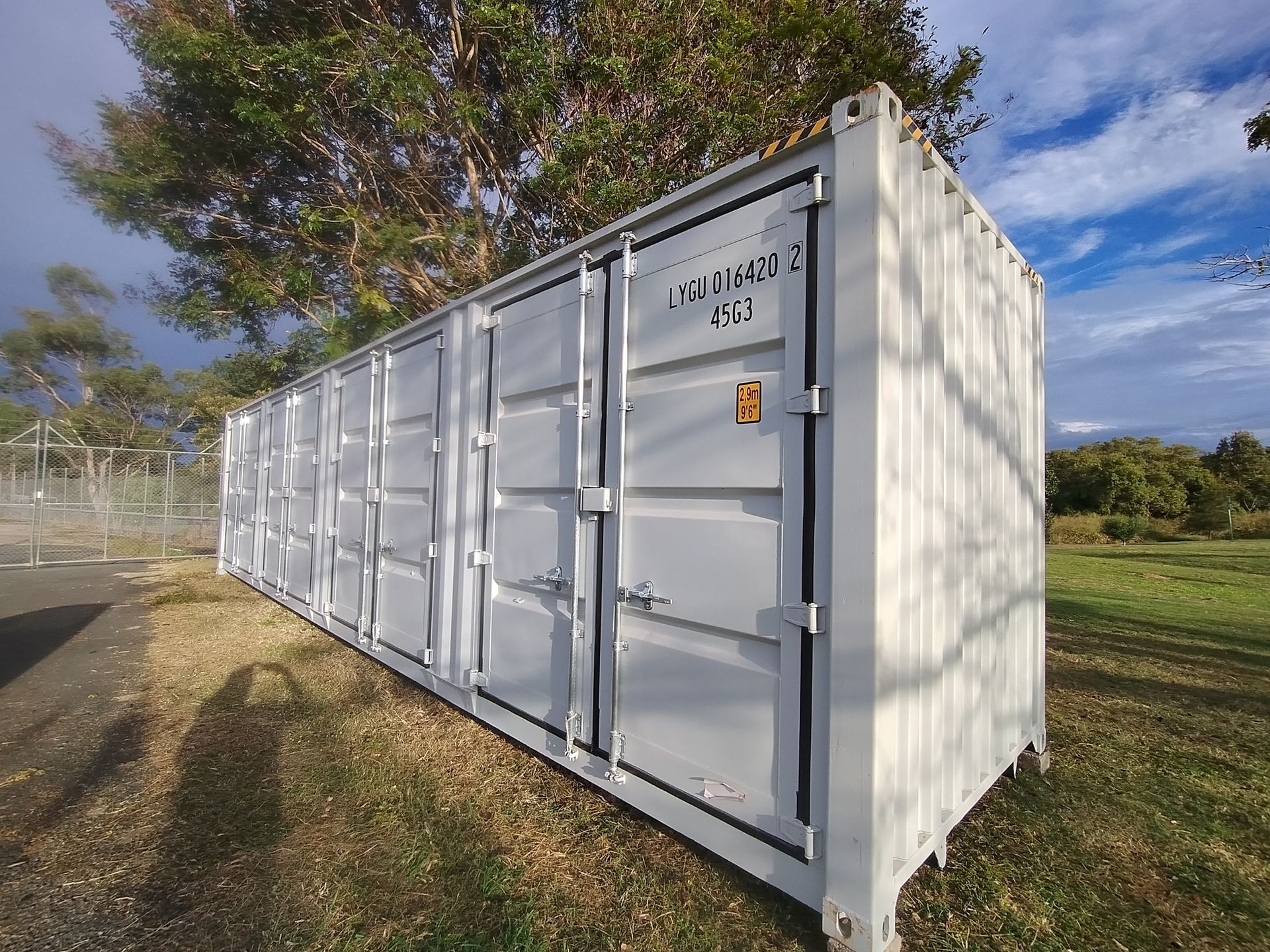 A white shipping container with multiple lockable storage doors positioned in a grassy field under a cloudy sky.