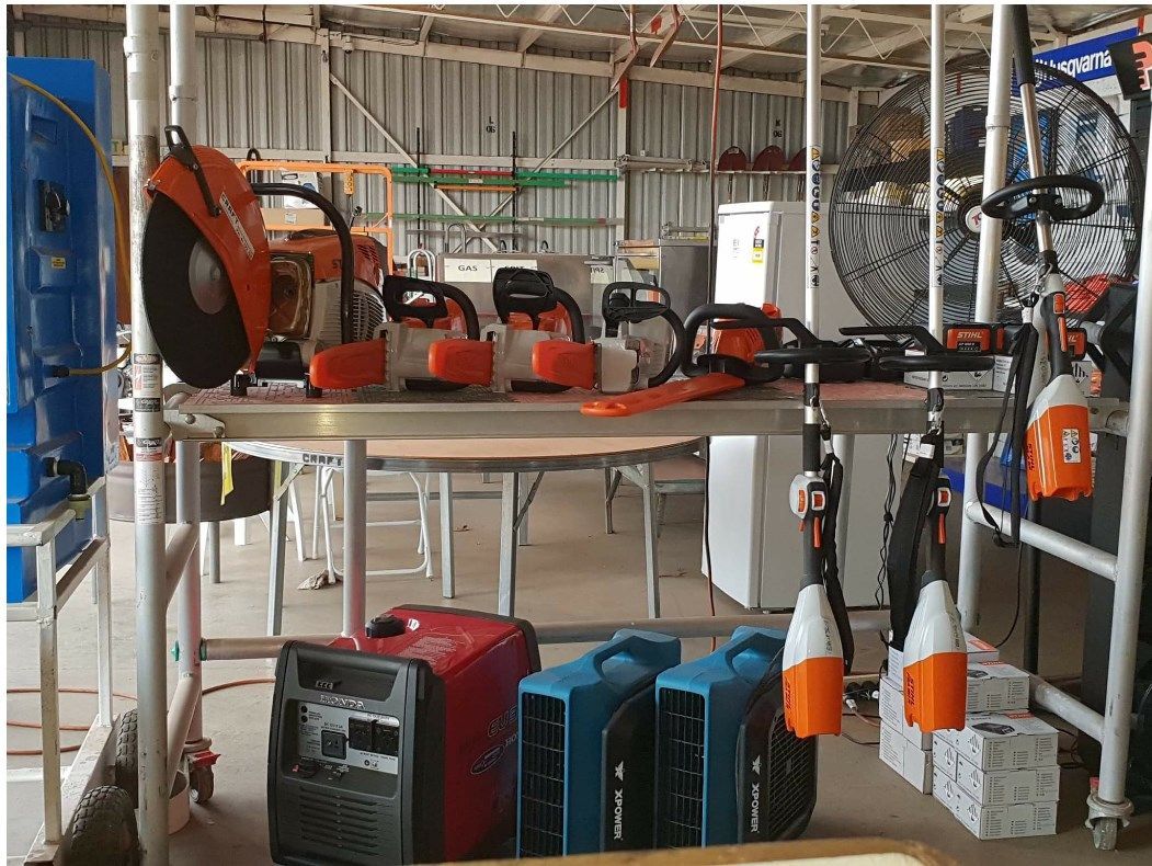A workbench displaying a cut-off saw, several chainsaws, and trimmers, with a generator and floor fans below.