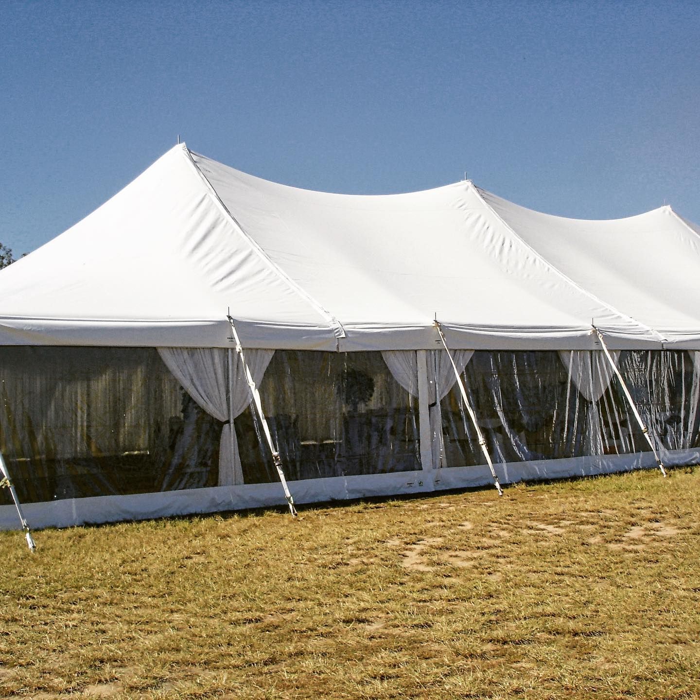 A white event tent with transparent side walls and draped curtains, set on a dry grassy field under a clear blue sky.