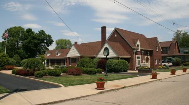 Brick building with brown roof, likely a funeral home, with landscaping, sidewalk, and blue sky.