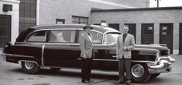 Two men standing next to a black hearse. The men wear suits and the setting appears to be outside a building.
