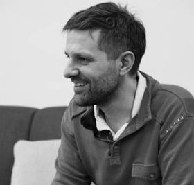 A man with a beard and sunglasses is smiling in a black and white photo.