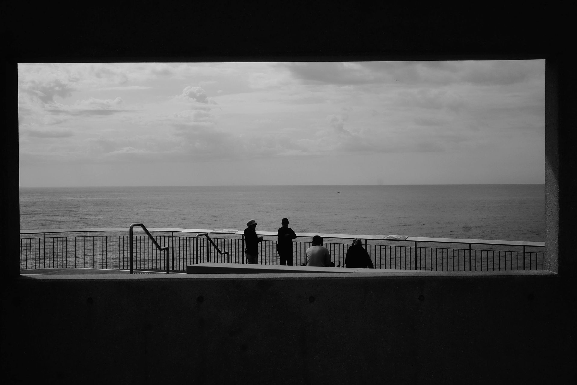 Three silhouetted figures on a seaside promenade, framed by a dark opening. The ocean and sky fill the background.
