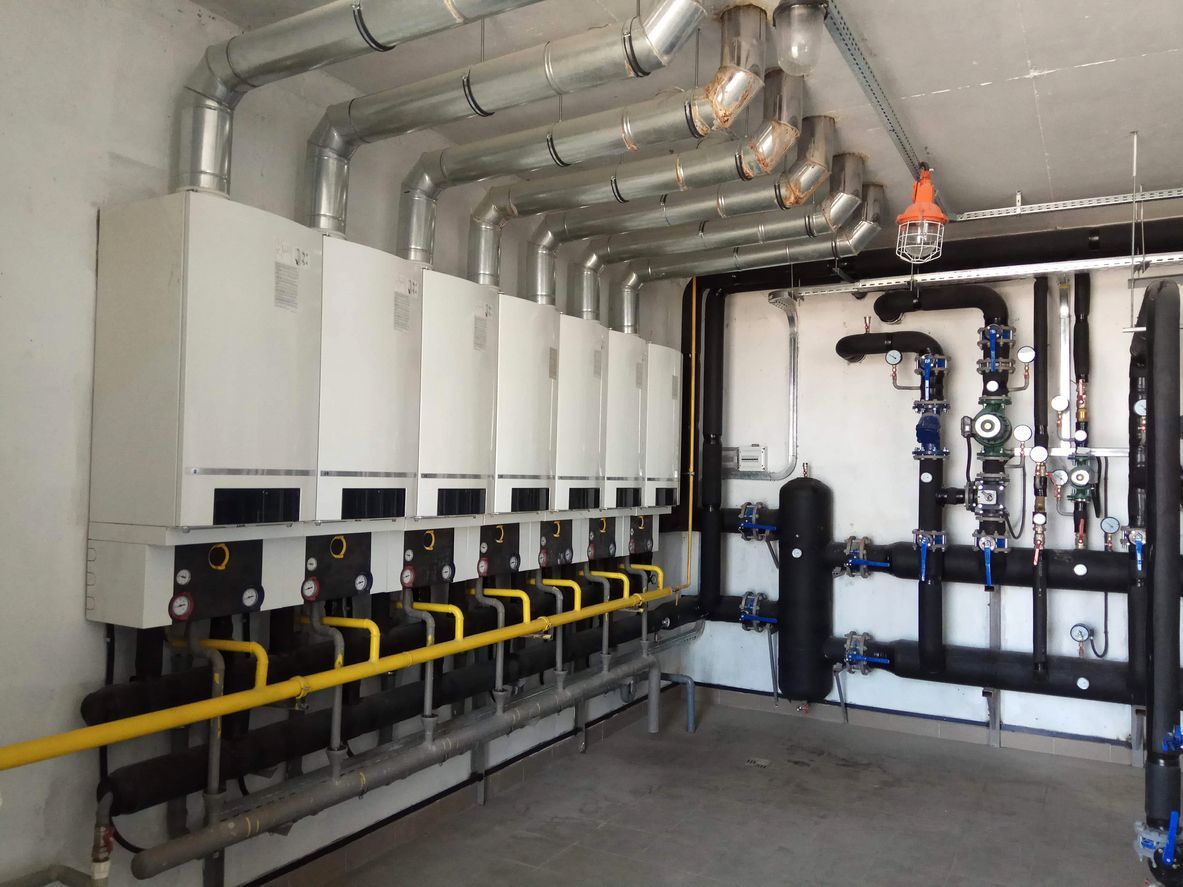 Mechanical room with wall-mounted boilers, pipes, and ductwork in a utility basement