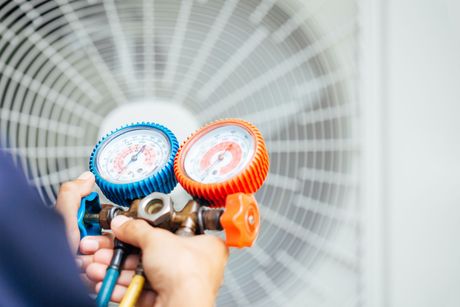 Hands holding a dual-gauge manifold with red and blue hoses in front of an outdoor AC unit fan