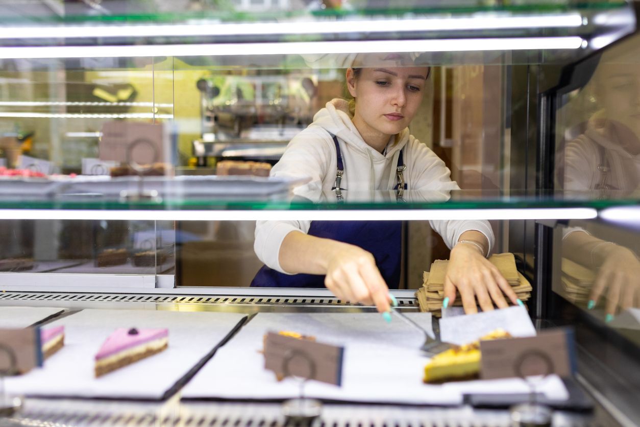 Bakery display case with a worker arranging cakes and pastries behind glass.