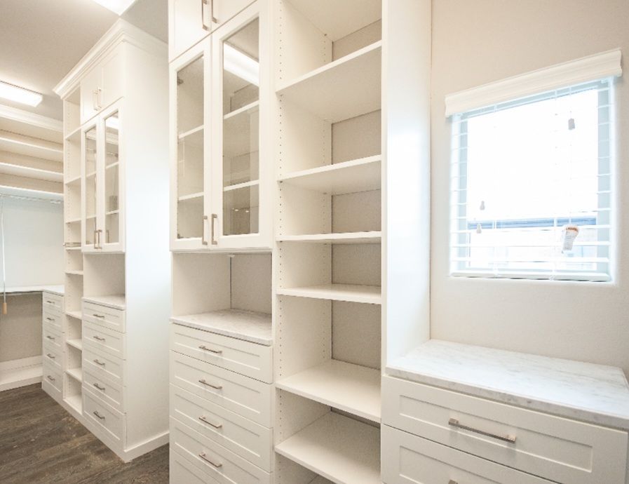 White custom closet with shelves, drawers, and glass-front cabinets; a small window with blinds is on the right.