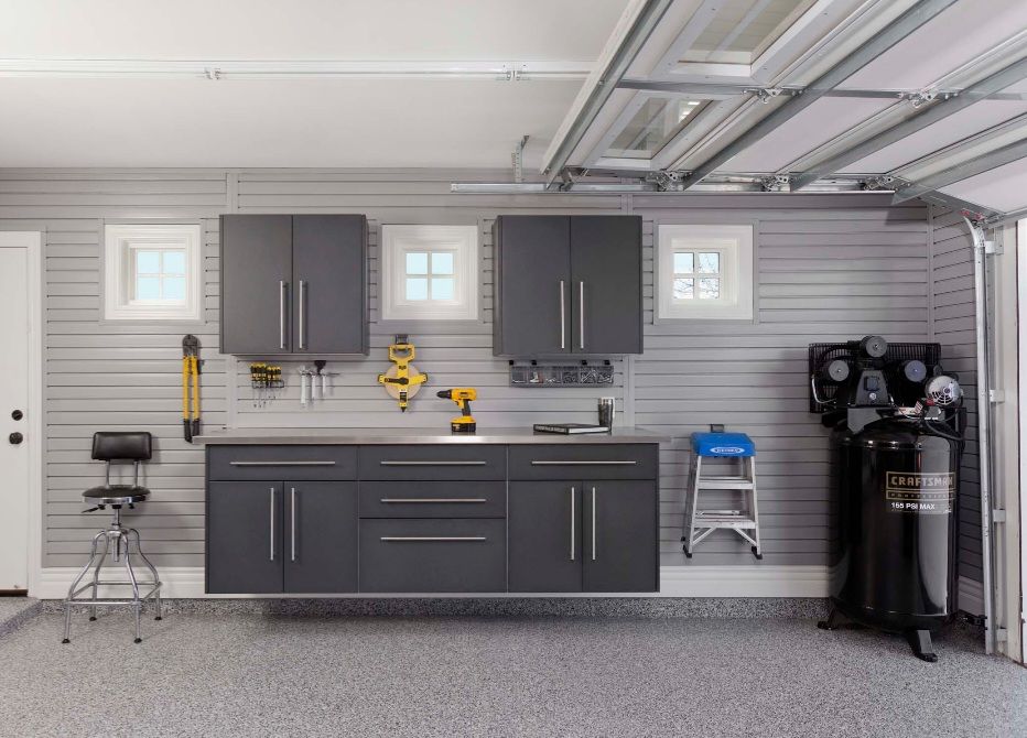 A garage with gray cabinets, tools, and a compressor. Epoxy floor, three windows, and the garage door is open.