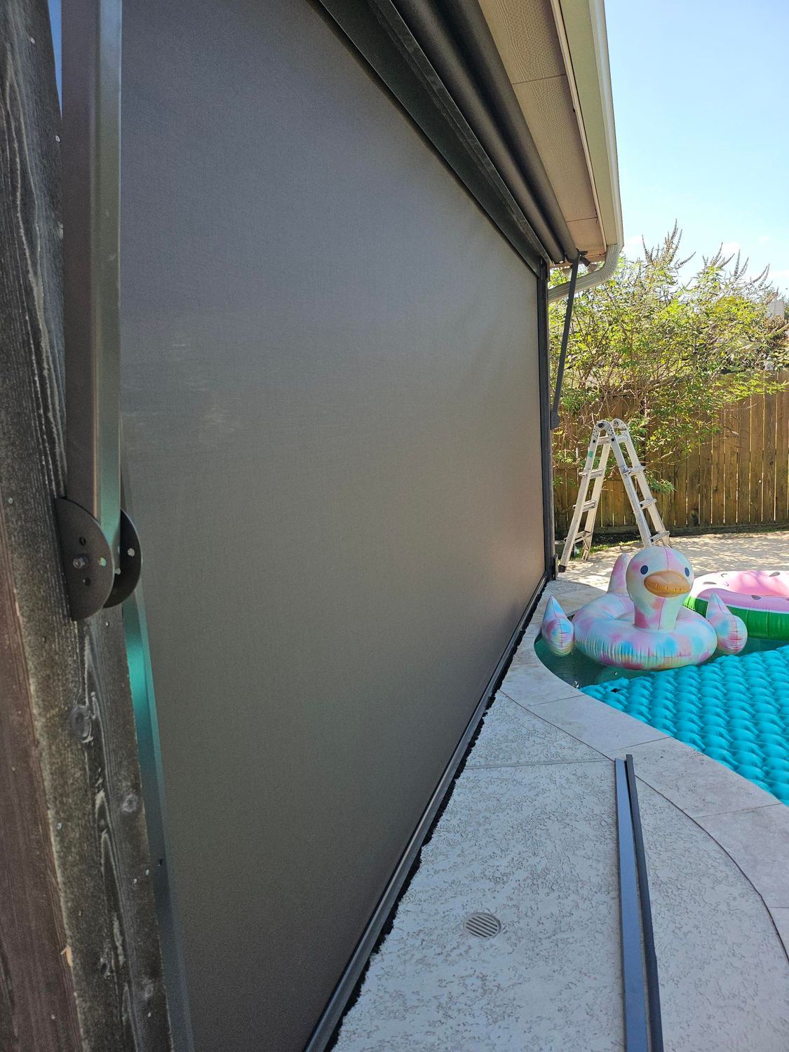 Exterior retractable sun shade next to a pool. Brown fabric, concrete patio, and a pool with inflatable toys.