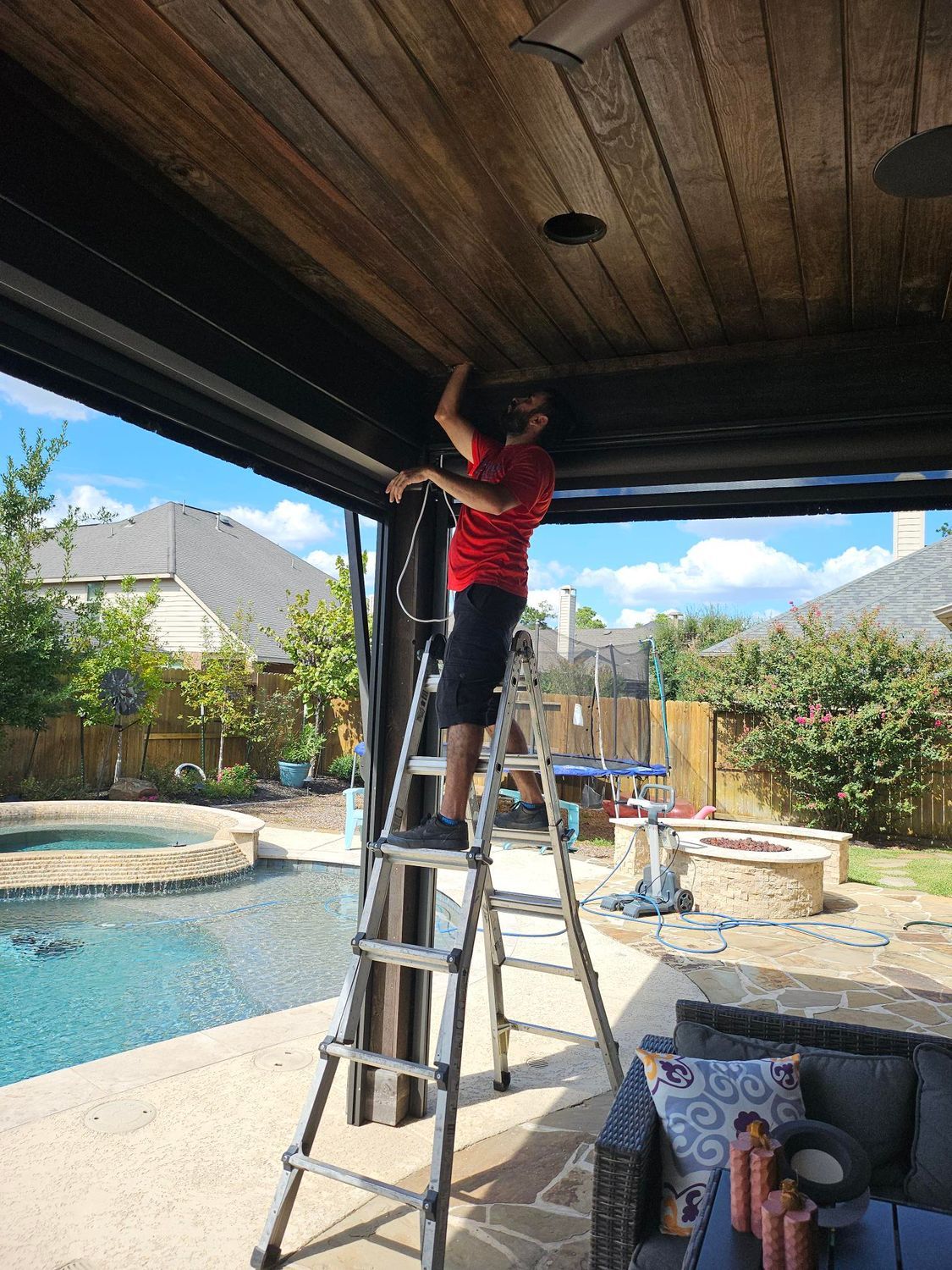 Man on ladder installing lights on a patio ceiling, pool and yard in the background.