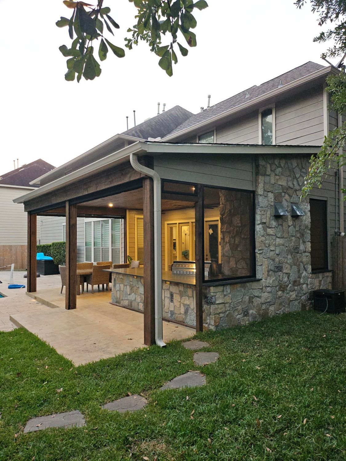 Covered outdoor patio with stone facade attached to a two-story house. Concrete patio, green lawn.