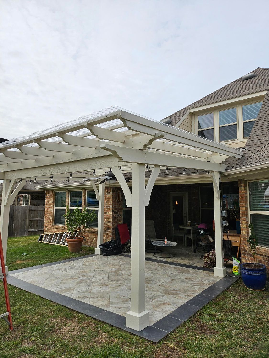 White pergola covers a patio, attached to a brick house. Overcast sky.